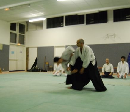 Photo floue, en noir et blanc, prise au dojo : deux pratiquants en mouvement sur le tatami ; au fond, d’autres pratiquants assis.