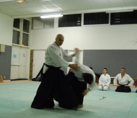 Un pratiquant en hakama accompagne son partenaire vers le sol ; derrière eux, d’autres élèves regardent, assis.